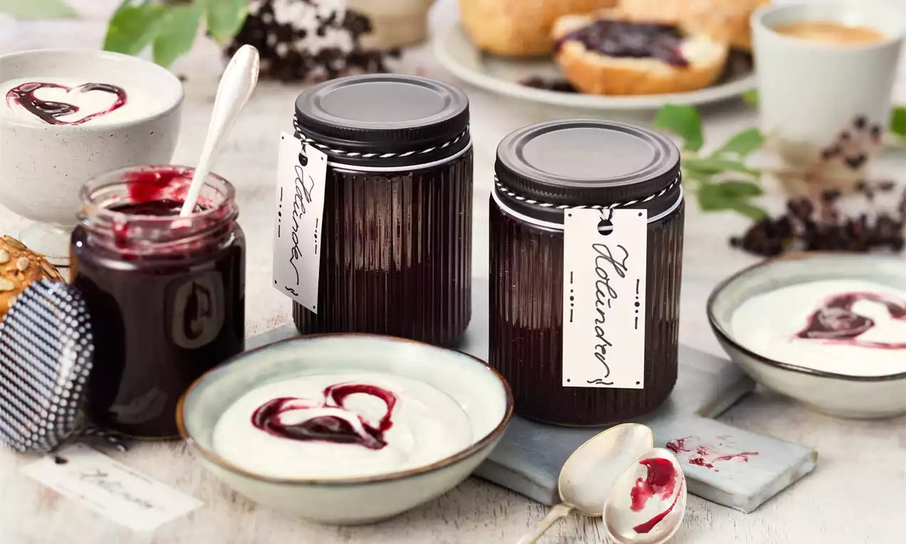 Dunkelrote Holundermarmelade in gerippten Gläsern mit weißem Etikett neben Joghurt mit Marmeladenherz und einem Löffel auf hellem Holz