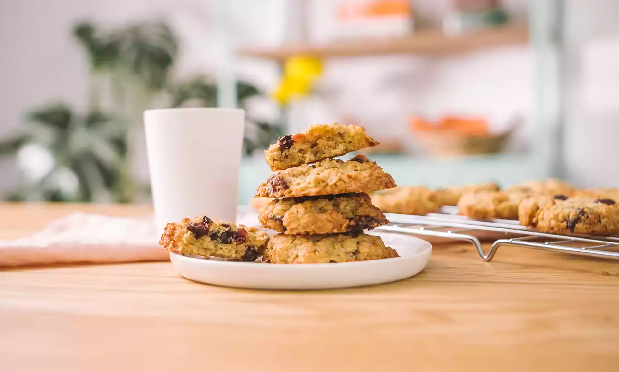 Galletas doradas de avena con bordes irregulares y textura rugosa apiladas en un plato blanco junto a una taza sobre mesa de madera clara
