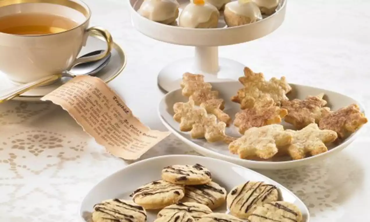 Verschiedene Sorten Teegebäck auf weißen Tellern, dekoriert mit Schokolade, Zuckerguss und Orangenzesten, neben einer Tasse Tee und Rezeptkarte