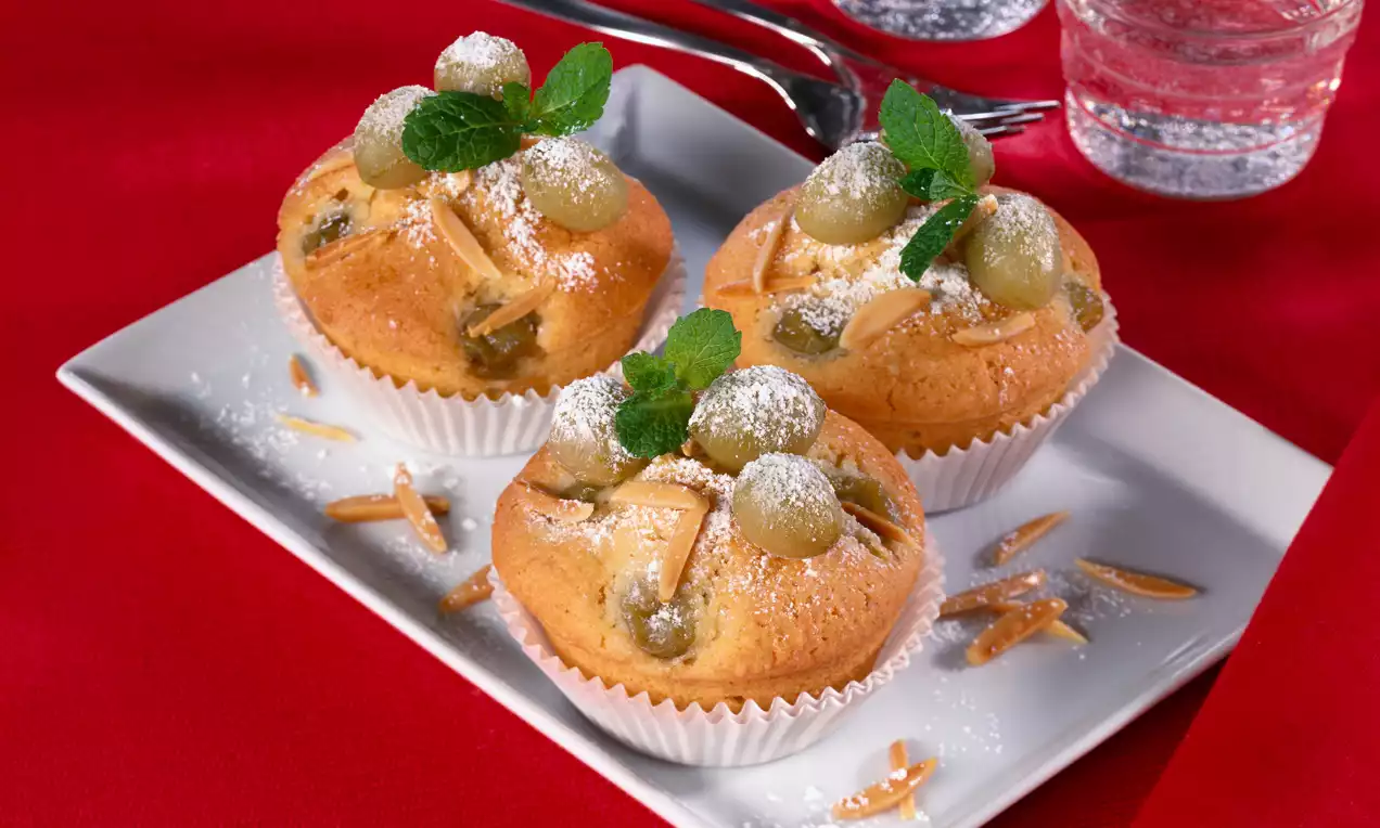 Goldene Stachelbeermuffins in Papierförmchen, garniert mit Mandelsplittern, frischen grünen Trauben, Minze und Puderzucker auf weißem Teller