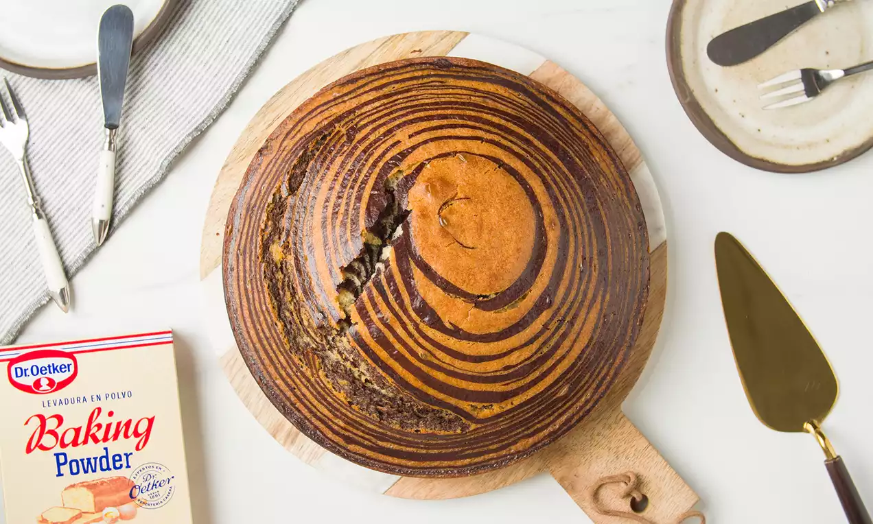 Bizcocho redondo sobre tabla de madera con líneas concéntricas marrón oscuro y doradas, corte en el centro mostrando su textura húmeda