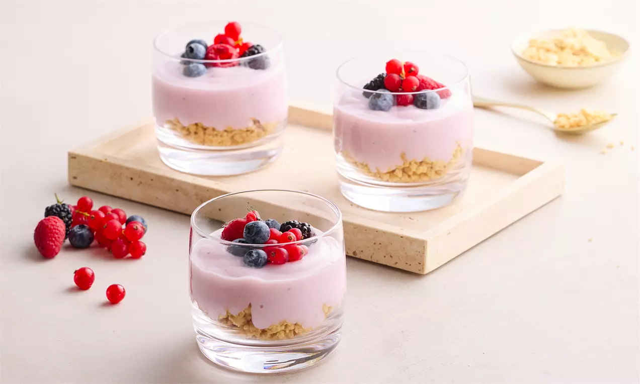 Şeffaf cam kup içinde kremsi pembe puding, altın rengi kıtır taban ve üstünde canlı kırmızı orman meyveleriyle zarif bir sunum