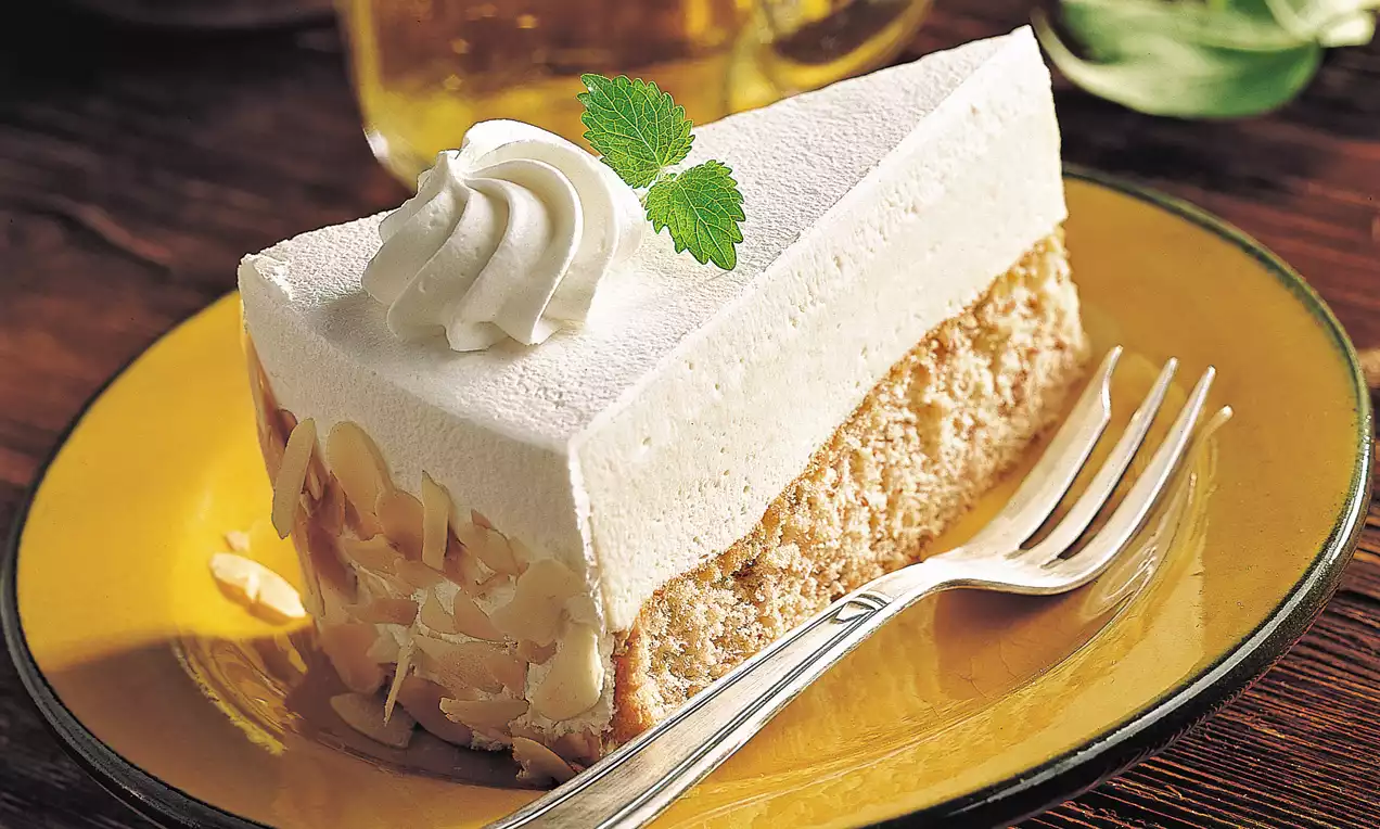 Part de gâteau crèmeux beige pâle, bordée d'amandes effilées, surmontée d'une rosace de chantilly et de feuilles de menthe sur une assiette jaune