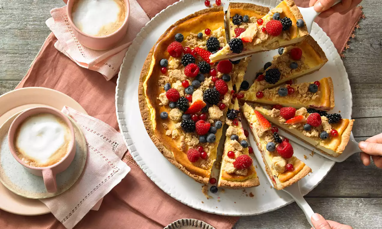 Skyr-Rahm-Kuchen mit goldbraunen Streuseln, frischen roten und dunklen Beeren auf weißer Platte neben Tassen mit Milchschaum