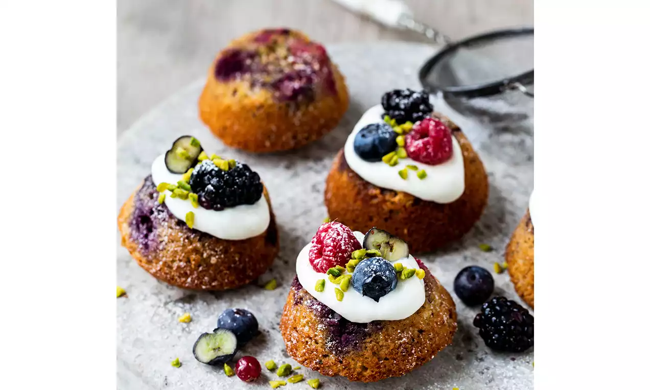 Muffins dorés garnis de crème blanche, baies colorées et éclats de pistache, posés sur une assiette saupoudrée de sucre glace