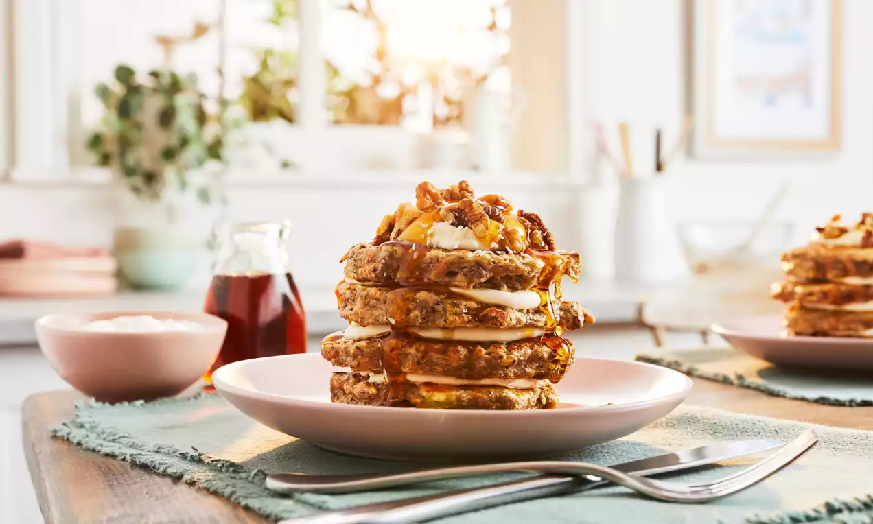 A stack of golden-brown carrot cake pancakes layered with cream cheese, topped with walnuts on a soft pink plate with a syrup jar nearby