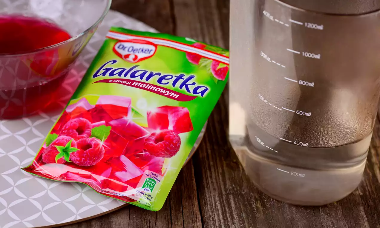 Opakowanie galaretki malinowej na białej podstawce obok szklanej miski z czerwoną galaretką i miarki z przezroczystą wodą