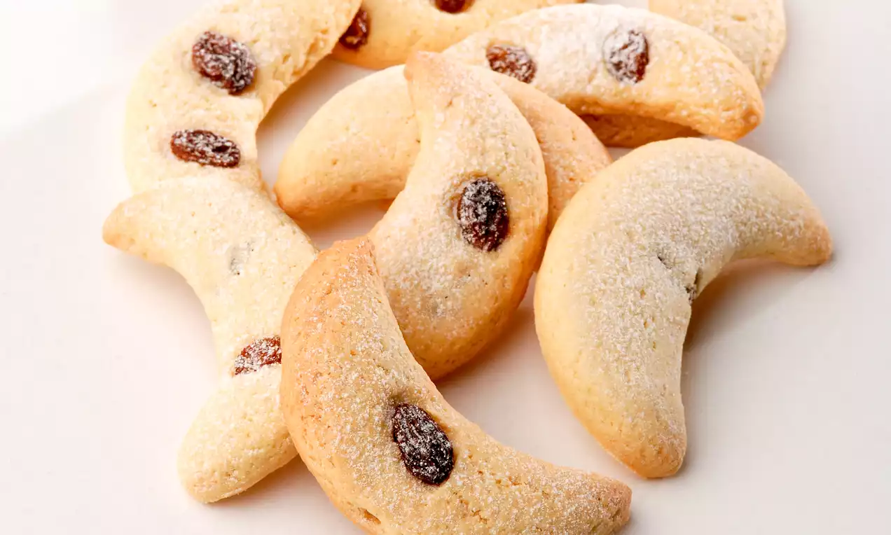 Altın sarısı ay şeklindeki kurabiyeler üzerlerine serpilmiş pudra şekeri ve parlak kuru üzümlerle sade bir tabakta sunulmuş