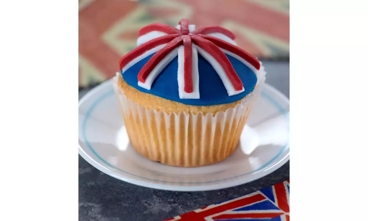 Golden cupcake with smooth blue icing, topped with precise red and white sugar stripes in Union Jack pattern on a white plate
