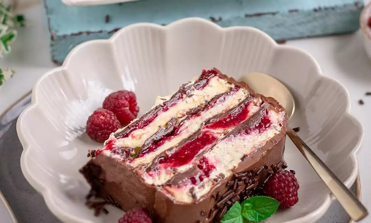 Komad slojevitog vanilija ledenog deserta s crvenim malinama i čokoladnim mrvicama na bijelom tanjiru, ukrašen svježim malinama i listićima mente