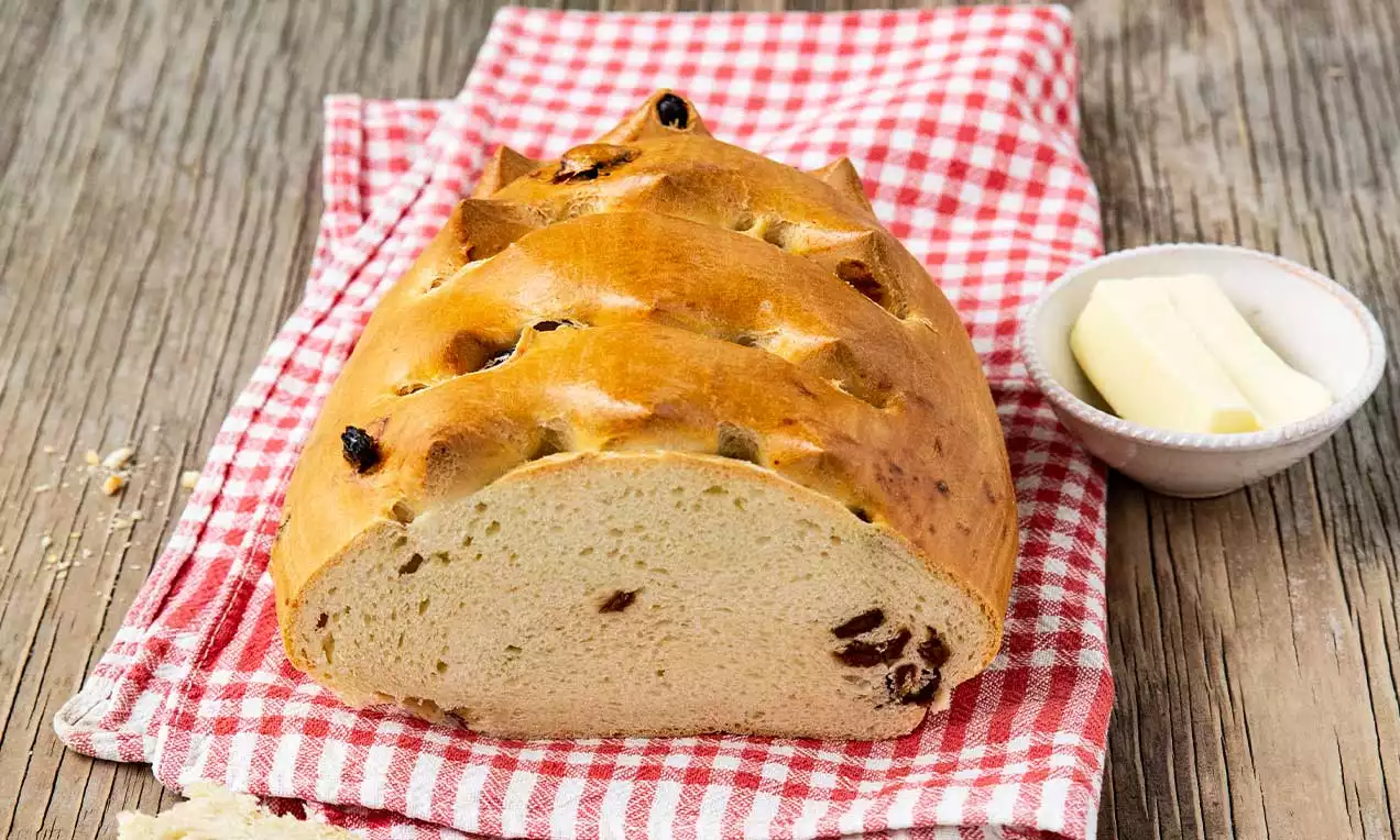Pain doré et moelleux parsemé de raisins secs, à la croûte entaillée, posé sur un tissu rouge à carreaux avec une coupelle de beurre