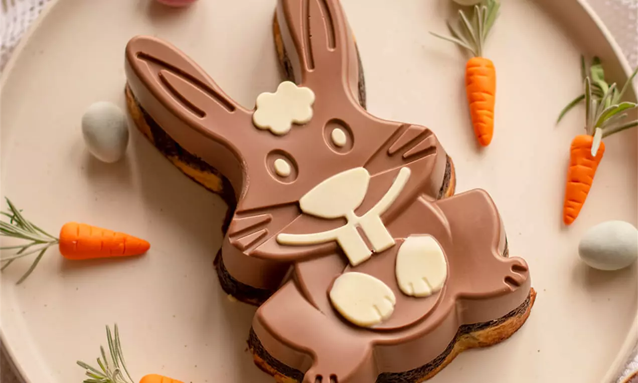 Čokoladni desert u obliku smeđeg zeca s bijelim detaljima, položen na tanjur, okružen šarenim jajima, malim mrkvama i grančicama ružmarina