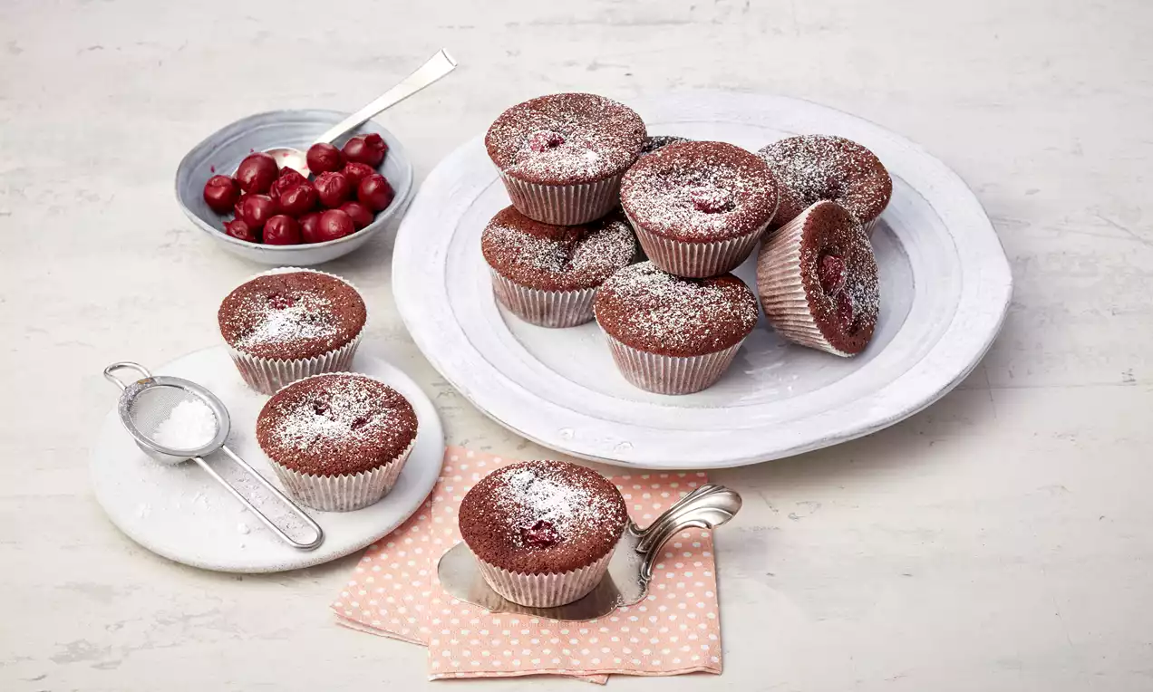 Schoko-Kirsch-Muffins mit dunkler Oberfläche, Puderzucker bestäubt, teils auf weißem Teller, Dekoration aus Kirschen, Sieb und rosa Tuch