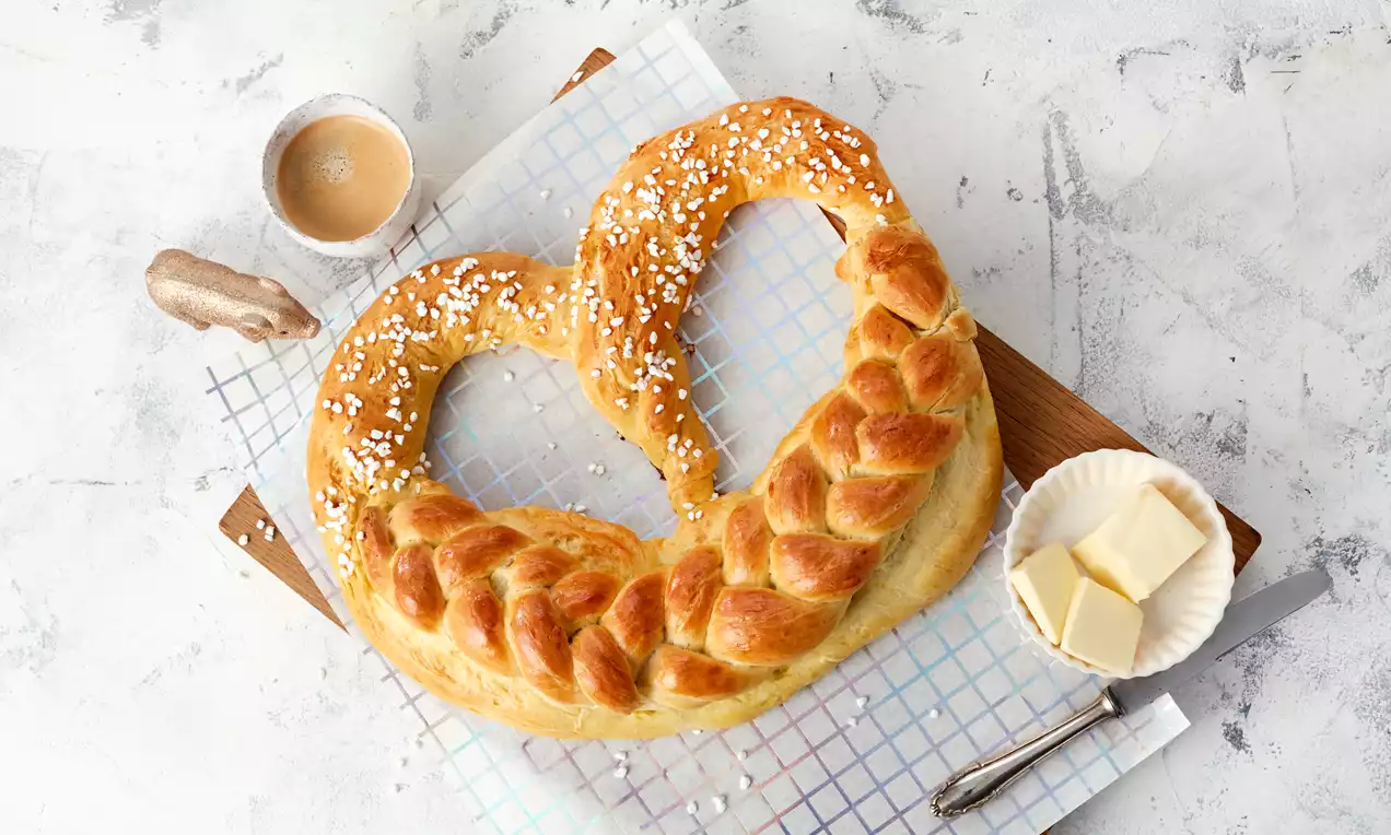 Goldbraune Neujahrsbrezel in Herzform mit glänzend gebackener Oberfläche und Zuckerperlen auf kariertem Backpapier mit Butter und Kaffee