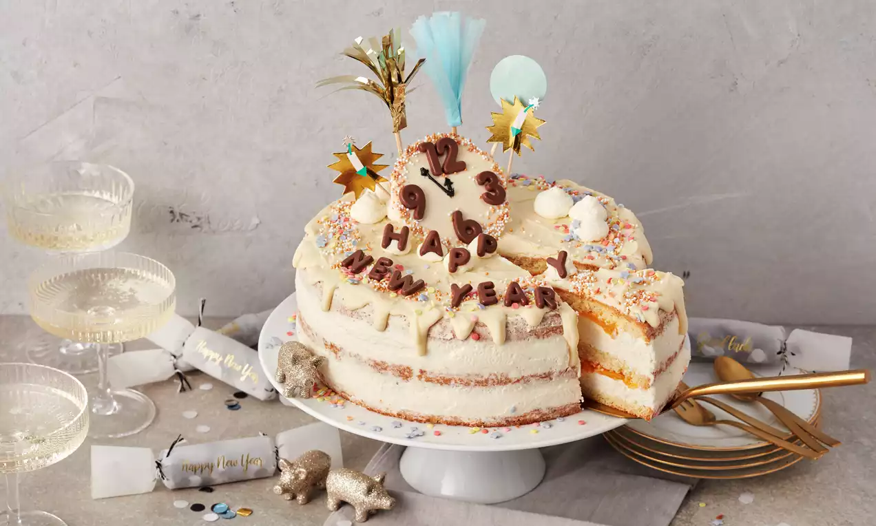 Eine helle, schichtweise gefüllte Silvester-Torte mit Creme, bunter Streudeko, Schokoladenuhr, Fondant-Schriftzug und Party-Toppern auf weißer Tortenplatte