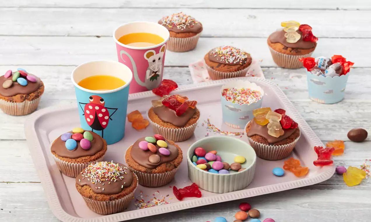 Bunte Geburtstagsmuffins mit Schokoladenglasur, dekoriert mit Zuckerstreuseln, Smarties und Gummibärchen auf einem rosa Tablett mit Saft in Bechern