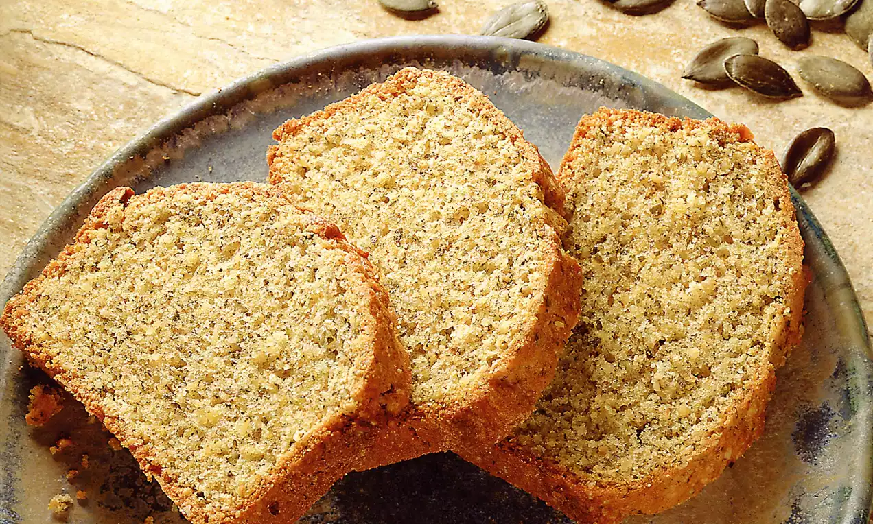 Drei goldbraune Kuchenstücke mit grober, körniger Textur auf dunklem Keramikteller, daneben lose Kürbiskerne auf hellem Untergrund