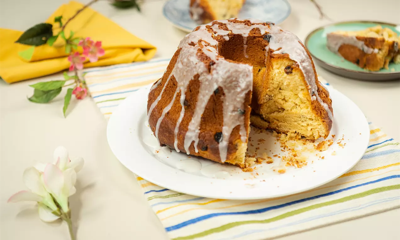Кръгъл козуначен кейк с топла златиста коричка, изрисуван с бяла захарна глазура, разрязан на голямо парче и поднесен върху бяла чиния