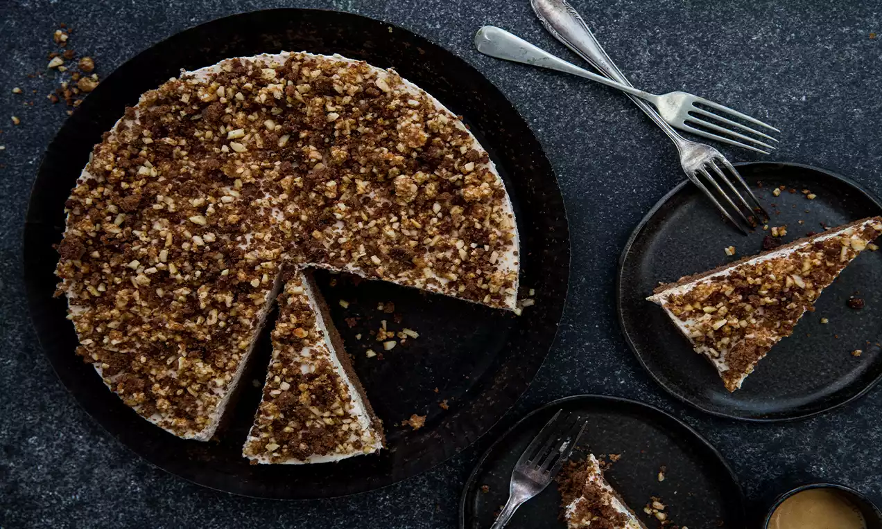 Runder Kaffeekuchen mit heller Creme und dunklem Streuselbelag, angeschnitten auf schwarzem Teller, daneben Kuchengabeln und Stücke auf kleinen Tellern