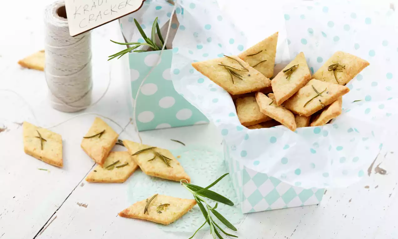 Zlato zapečeni, rombasto oblikovani krekerji z zelišči, posuti z drobnimi vejicami rožmarina, razporejeni v pastelno zeleni škatli s pikami