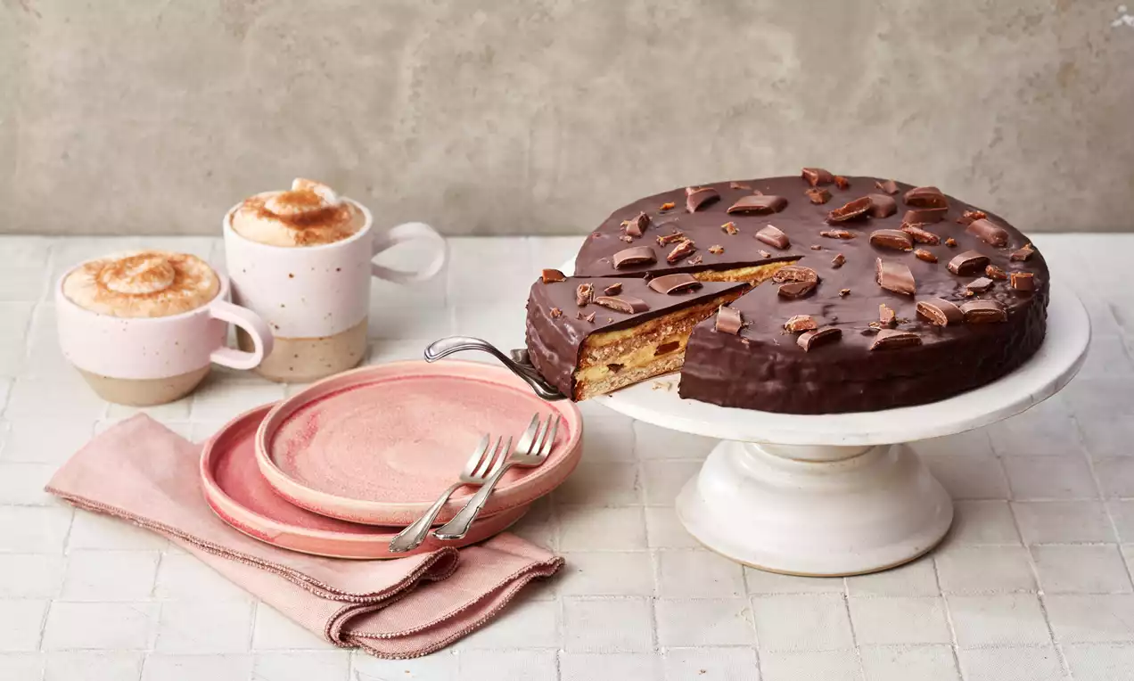 Runde Daim®-Torte mit glänzender Schokoladenschicht, bestreut mit Karamellstücken, angeschnitten auf weißer Tortenplatte, daneben rosa Teller und zwei Cappuccino-Tassen