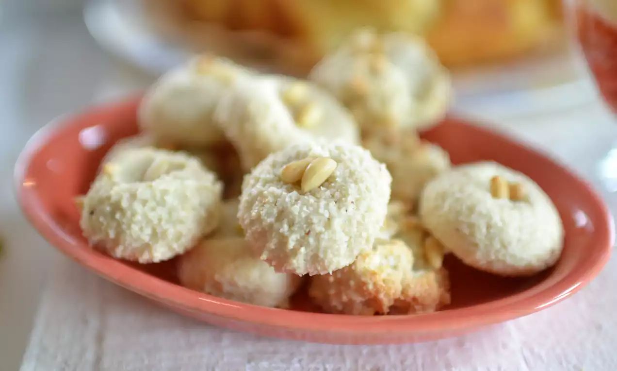 Kleine ronde witte koekjes met een kruimelige textuur, gegarneerd met pijnboompitten, op een lichtroze schaaltje geplaatst