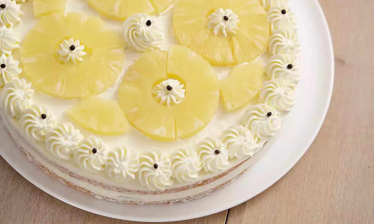 Tort rotund decorat cu felii aurii de ananas și moturi delicate de frișcă albă, pe un platou simplu alb plasat pe lemn deschis