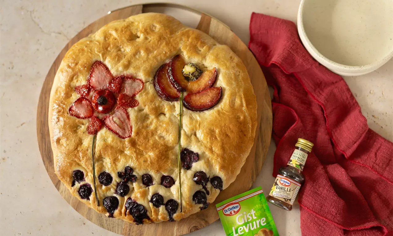 Focaccia dorée ornée de motifs floraux en fruits rouges et pruneaux, sur une planche en bois avec des accessoires rouges et beige