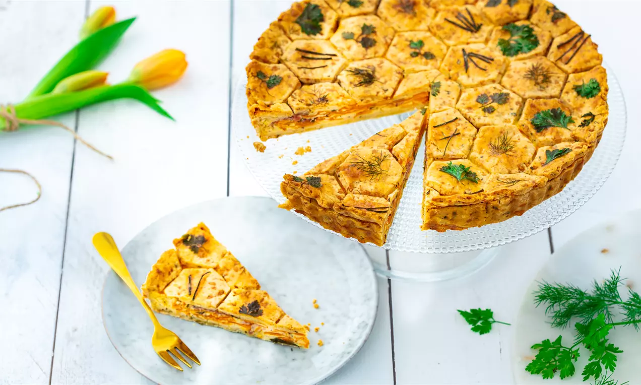Златисто-кафяв солен тарт с шестостенни форми, украсен с морковени пластове и зелени билки върху крехко тесто, сервиран на стъклена стойка