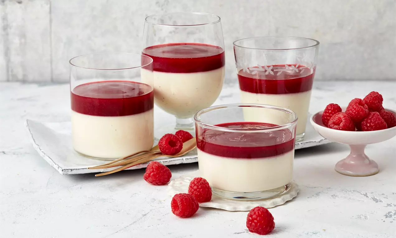 Panna cotta in Gläsern mit glänzend roter Himbeersauce, umgeben von frischen Himbeeren auf weißem Untergrund und einer kleinen Schale