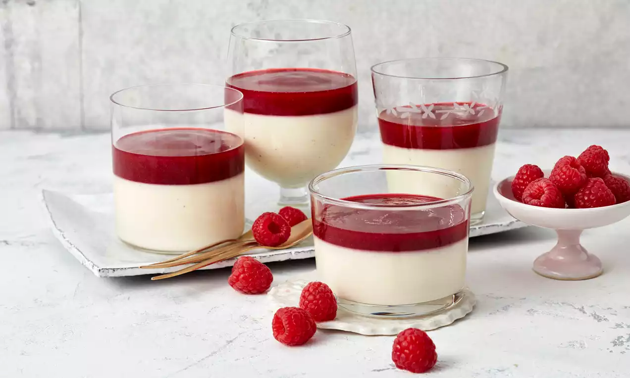 Weiße, cremige Panna Cotta in Gläsern mit glänzend rotem Himbeerspiegel, dekoriert mit frischen Himbeeren auf einem hellen Untergrund
