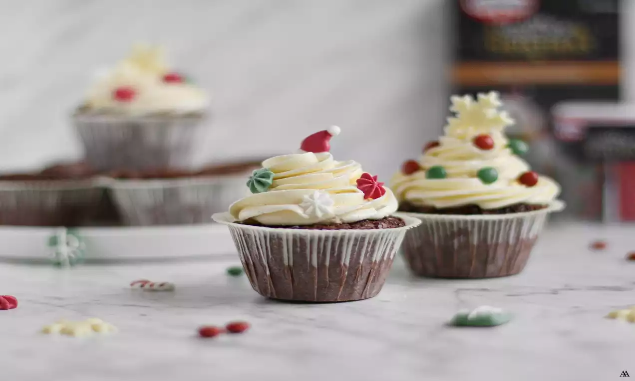 Chokladmuffins med krusig vit marängsmörkräm, dekorerade med färgglada julmotiv, placerade på marmorbord med strödda godisbitar