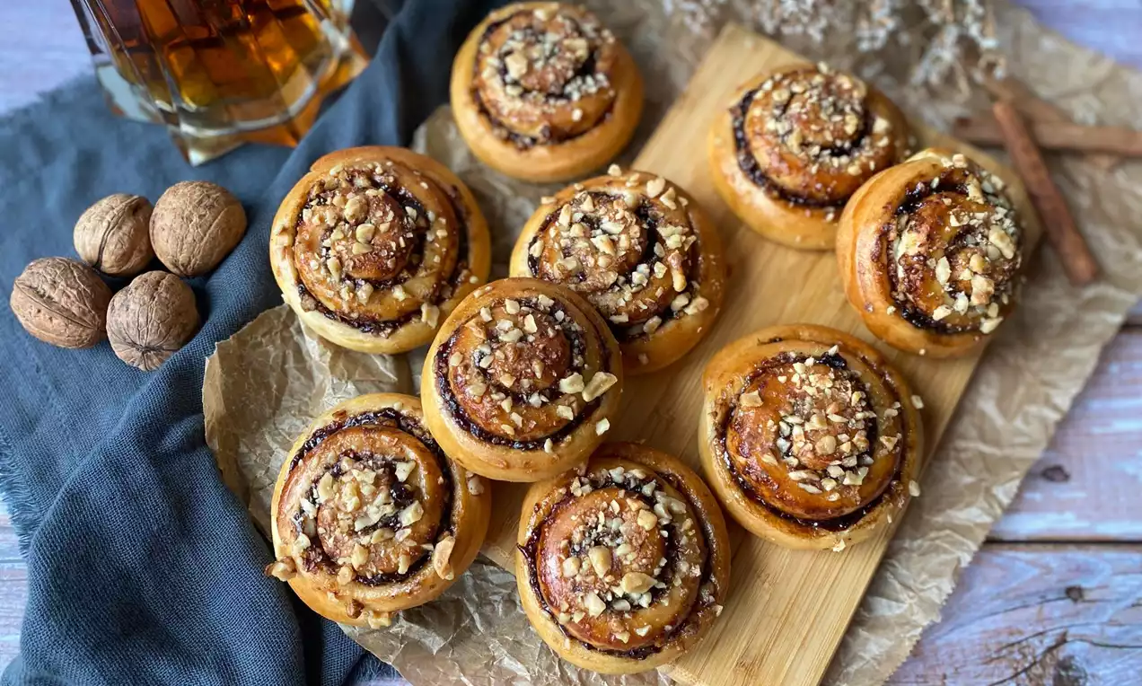 Zlatavé povidlové šneky s nádechem skořice, posypané jemně mletými ořechy, uspořádané na tmavé látce vedle celých vlašských ořechů