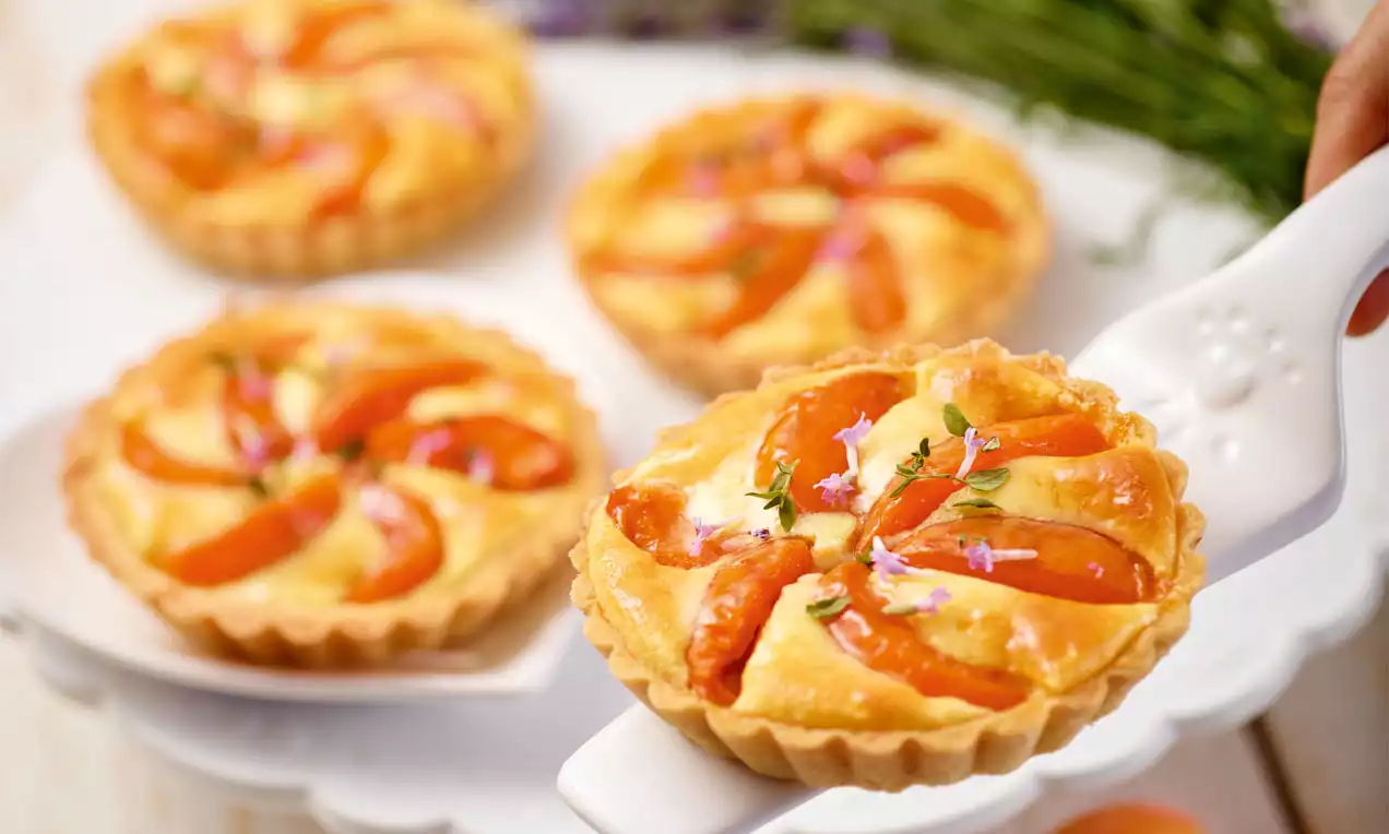 Goldene Marillen-Tartelettes mit knusprigem Rand, belegt mit saftigen Aprilschnitten, Thymian und Lavendelblüten auf weißem Teller