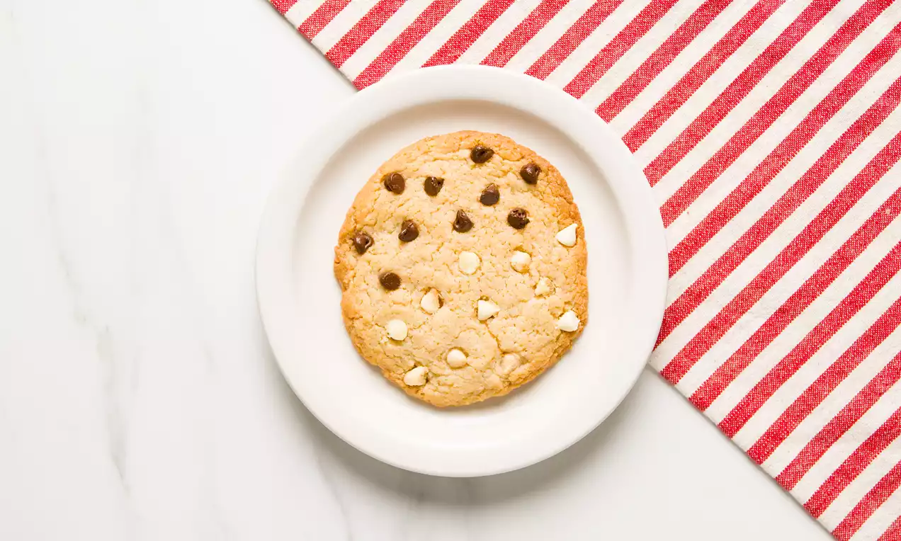Galleta redonda y dorada con chips de chocolate blanco y oscuro sobre un plato blanco, junto a un mantel de rayas rojas y blancas