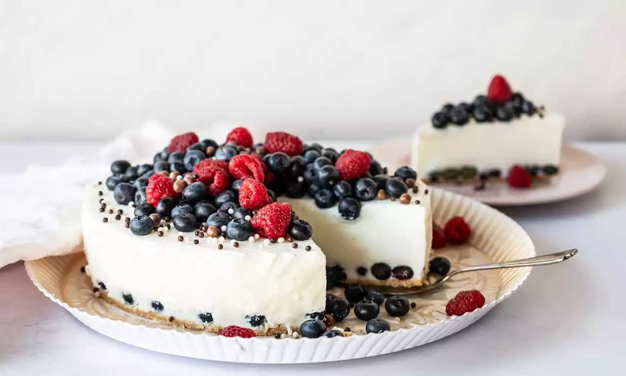 Weiße Quarktorte mit glatter Oberfläche, dicht mit blauen Heidelbeeren, frischen Himbeeren und Schokokrümeln bedeckt auf einem hellen Teller