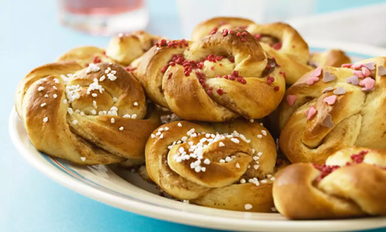 Kanelboller med gyllenbrun overflate og perlesukker, rosa strøssel og hjerter samlet på et hvitt serveringsfat mot blå bakgrunn