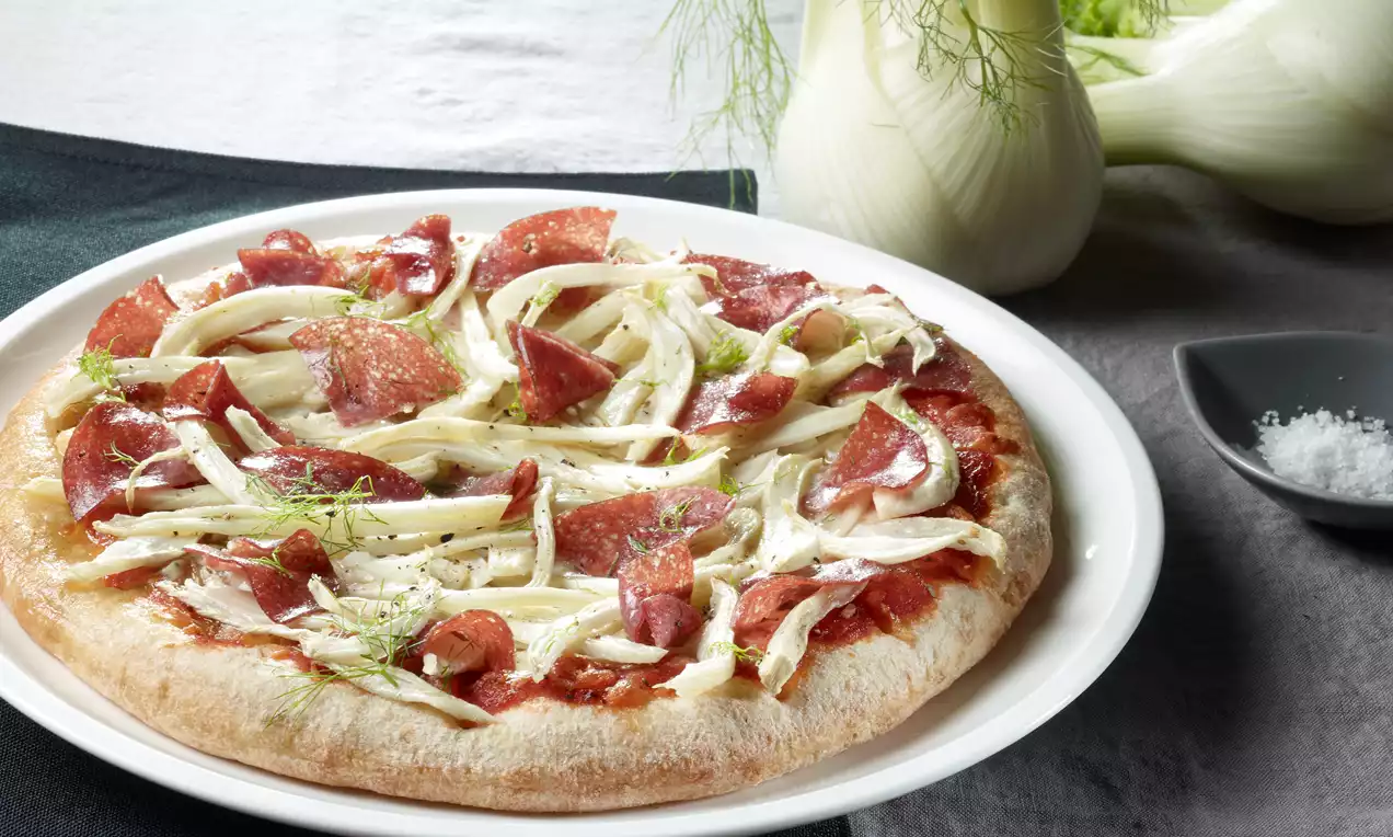 Runde Pizza mit goldbraunem Boden, belegt mit roten Salami-Stücken, weißen Fenchelstreifen und grünen Fenchelspitzen auf weißem Teller