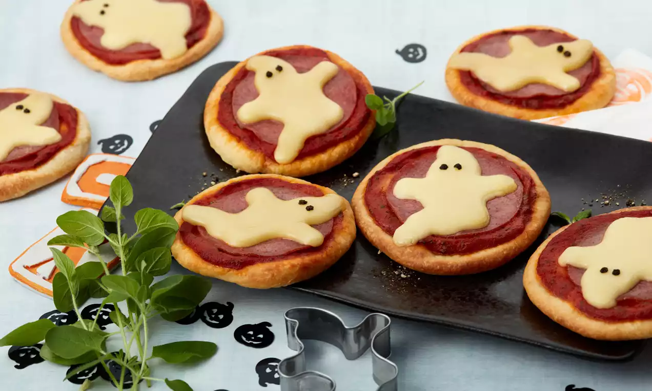 Kleine ronde pizzinis met rode tomatenlaag, bedekt met lichtgele kaas in spookvorm op een zwarte schaal, omlijst door kruiden en decoraties