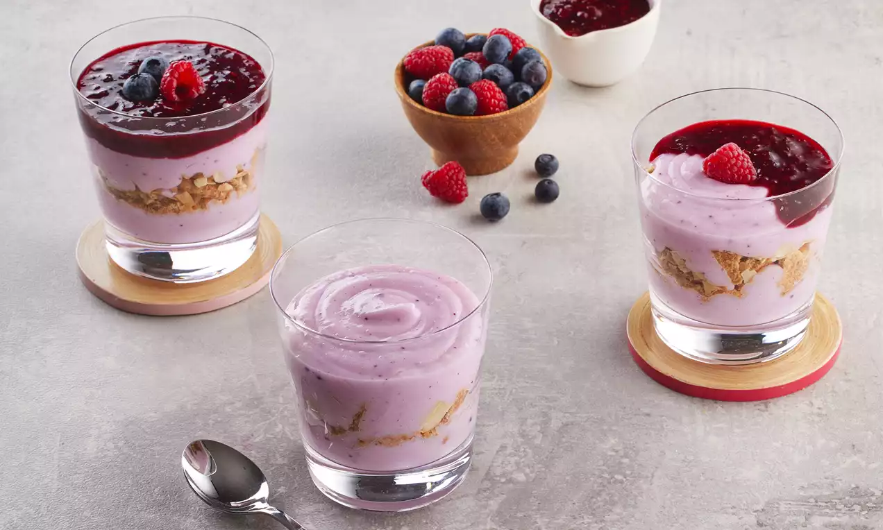 Küçük cam kupalarda kat kat dizilmiş mor kremalı tatlı, üzerinde böğürtlen sosu ve frambuaz taneleriyle sade bir sunum