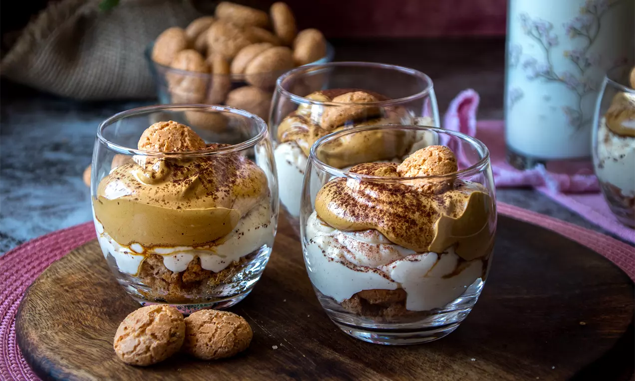 Слоесто фрапе тирамису в стъклени чаши с кремообразна текстура, поръсено с какао и гарнирано с амарети върху дървена дъска