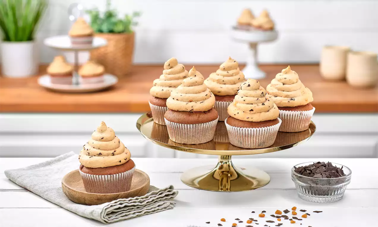 Schoko-Karamell-Cupcakes mit goldbrauner Cremehaube und Schokostückchen auf einer goldenen Tortenplatte, dekoriert mit gestreuten Karamellstücken