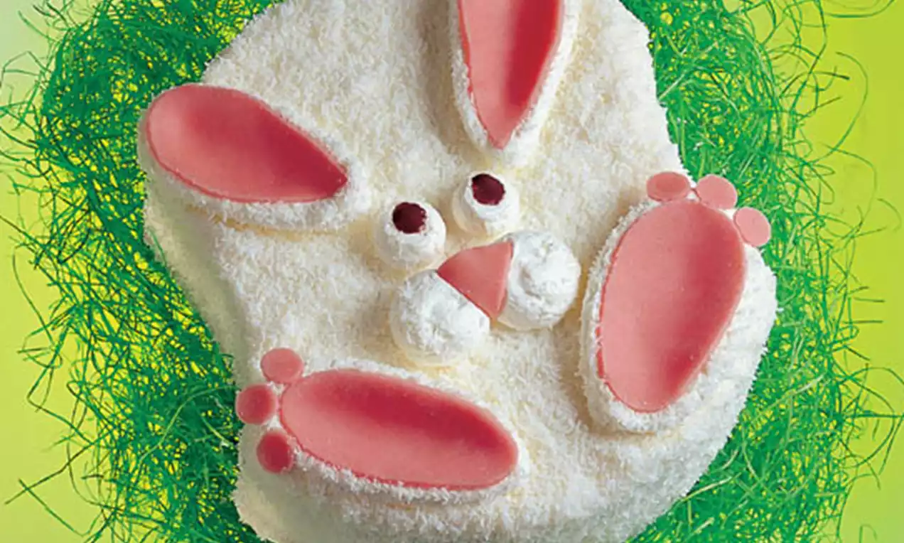 Witte paashaasvormige taart met roze oren en poten, bolle textuur bedekt met kokos, liggend op felgroen decoratief gras