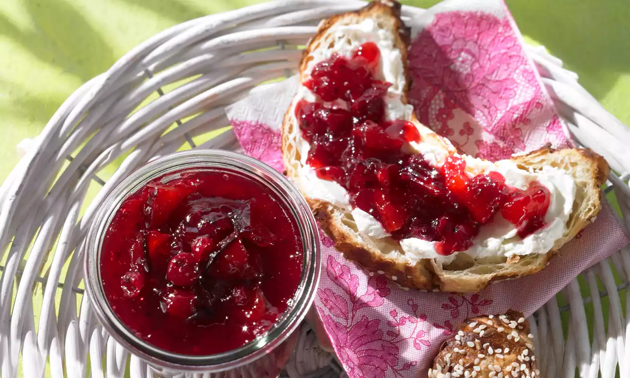 Confiture rouge rubis aux morceaux de fruits brillants servie dans un pot en verre et étalée sur une tranche de pain doré avec du cream cheese