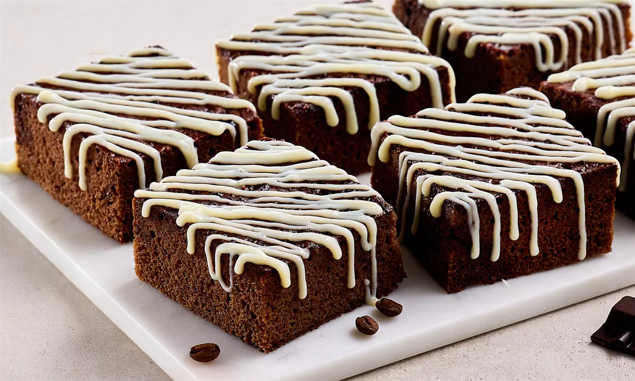 Kahverengi nemli kek dilimleri, üstünde beyaz ve kahverengi sos şeritleriyle sade bir tabakta kahve çekirdekleriyle sunulmuş