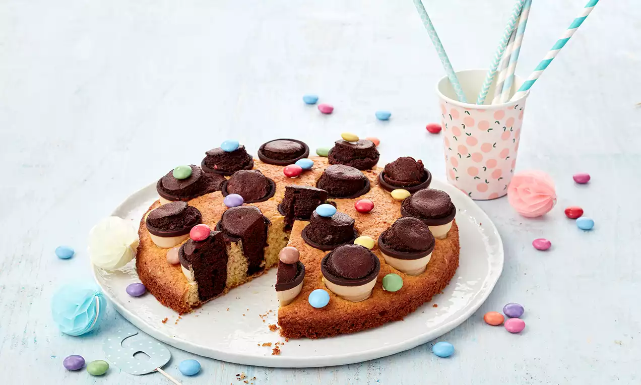 Runder, goldbrauner Kuchen mit bunten Schokolinsen, Mini-Brownies und Schokokeksen, auf weißer Platte mit Party-Dekoration arrangiert