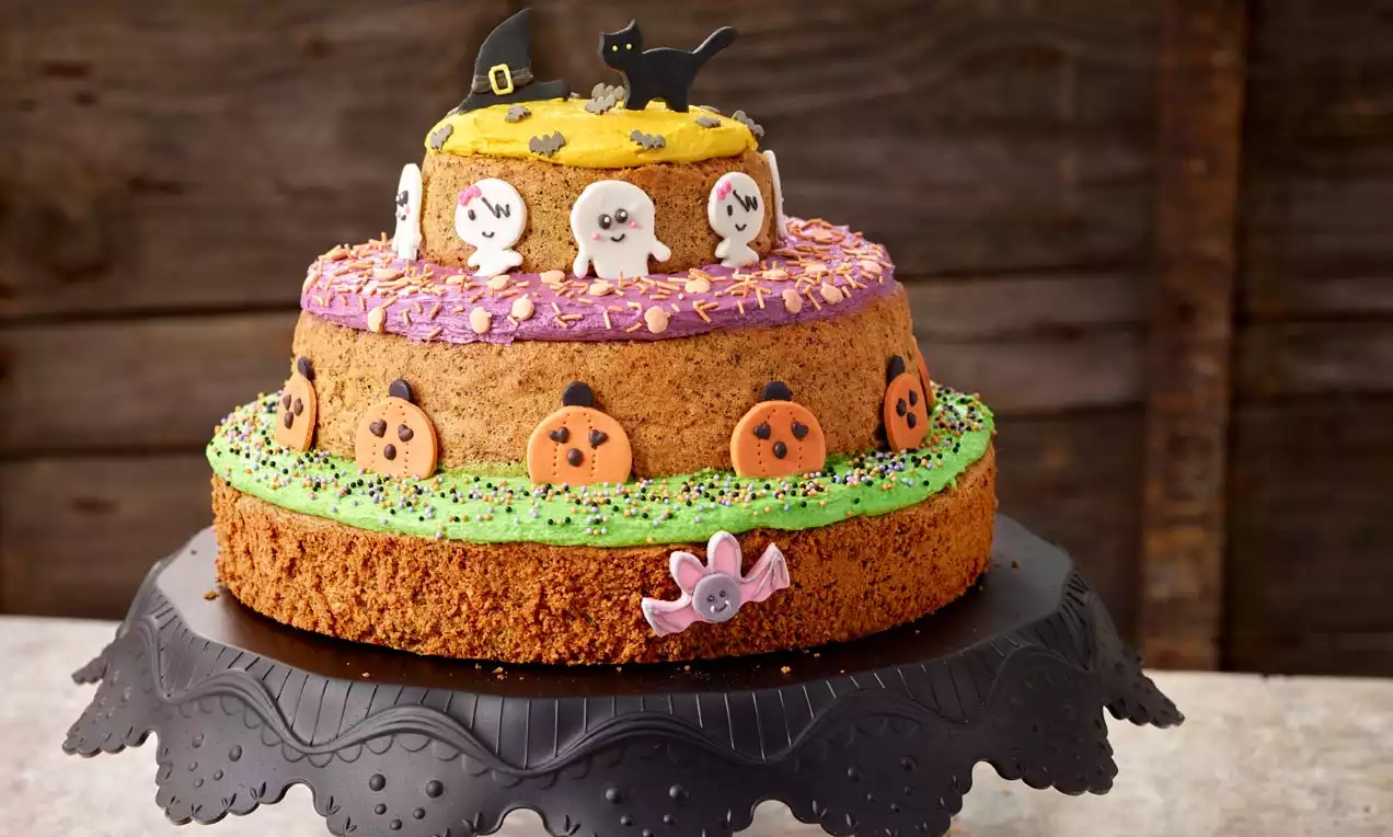 Zweistöckiger Naked Cake mit orange-braunen Böden, dekoriert mit Geistern, Kürbissen, grünem und lilafarbenem Zuckerguss auf schwarzer Tortenplatte