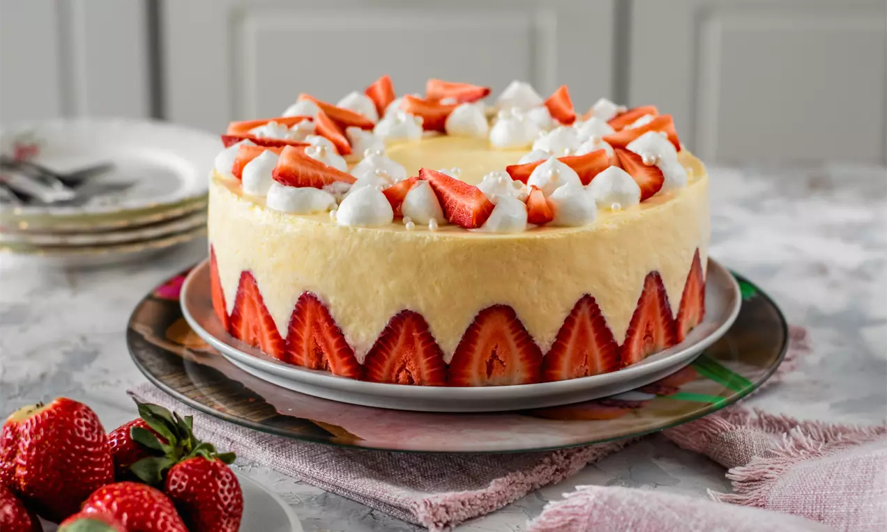Кръгла торта с кремообразен бледожълт баварски крем, обгърната с ягодови резени и декорирана с цели ягоди и пухкави бели розички върху шарена чиния