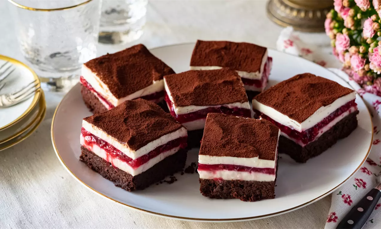 Квадратни парчета с тъмнокафяв шоколадов блат, бял слой чийзкейк и яркочервена вишнева глазура, нежно поръсени с какао върху бяла чиния