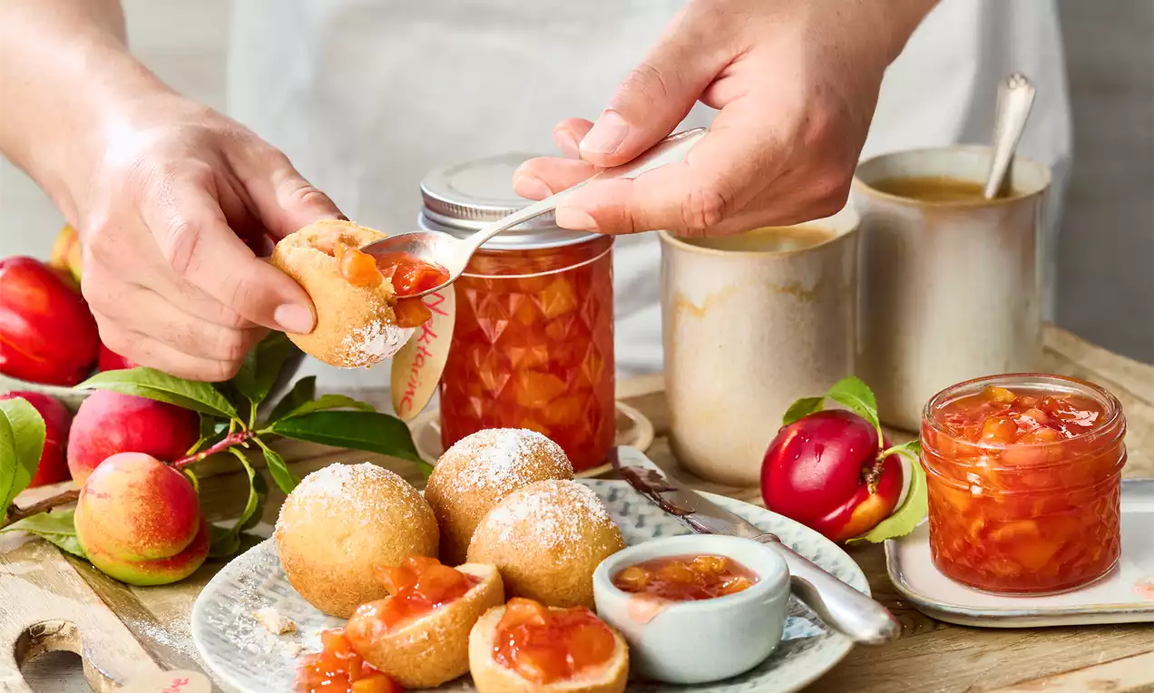 Nektarinenmarmelade in einem gläsernen Konfitürenglas mit leuchtend orangefarbener Fruchtstückchen, serviert mit Gebäck und frischen Nektarinen